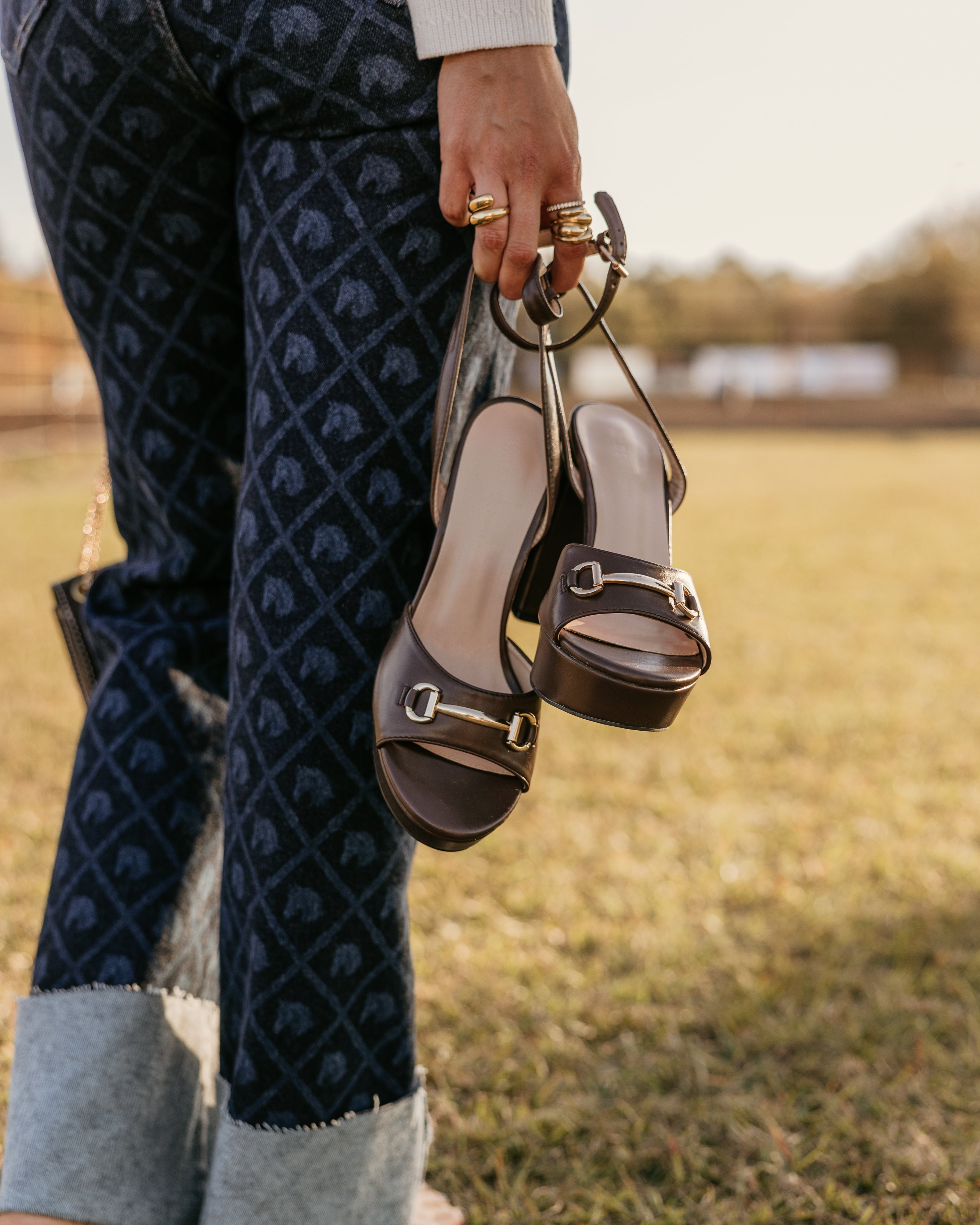 Woman holding horsebit accessory heeled sandals outdoors, wearing patterned wide cuff jeans