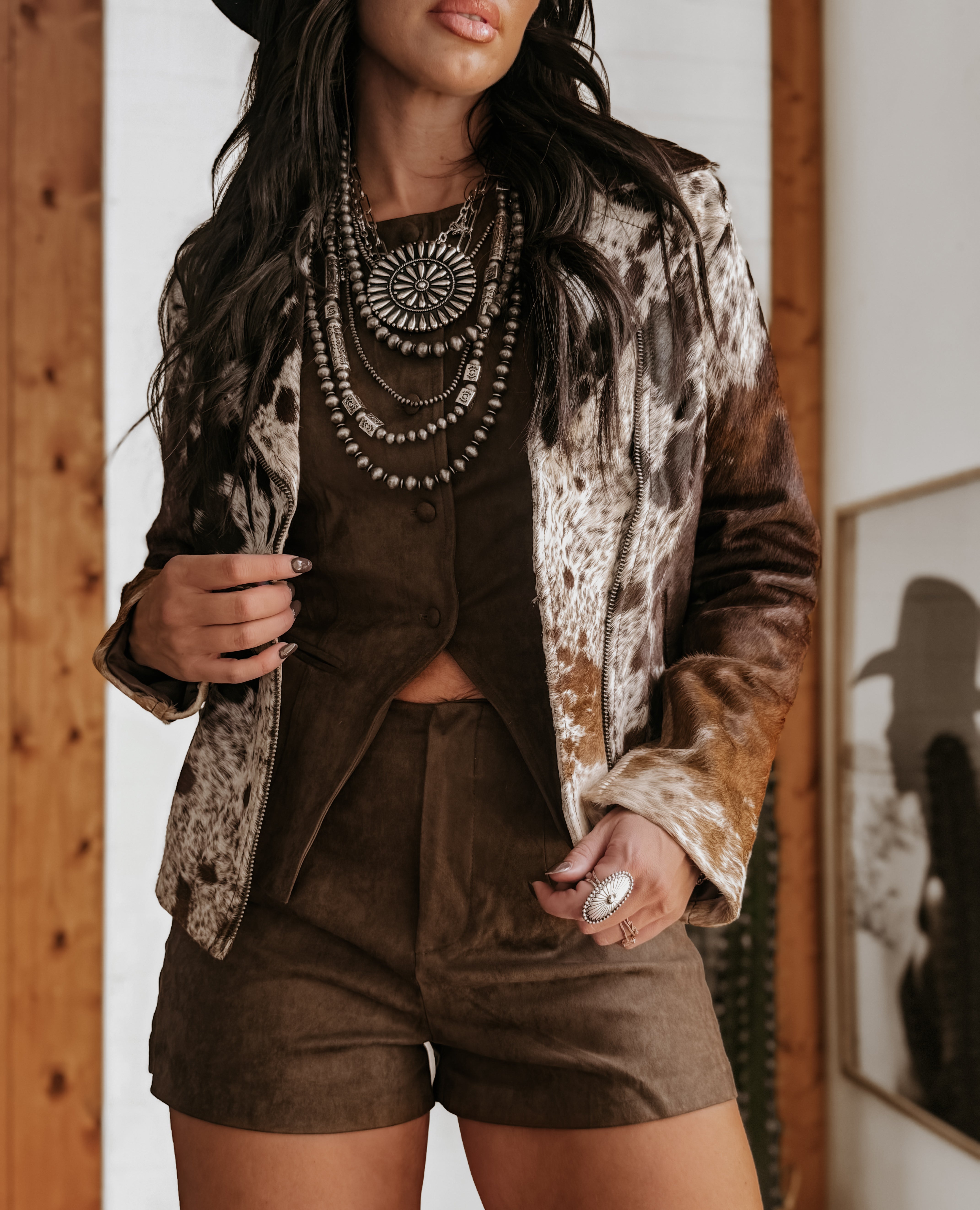 Woman in a western cow print jacket, brown suede shorts, and layered boho necklaces.