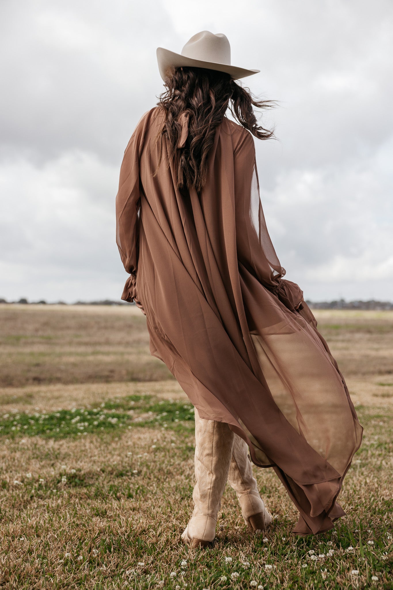 Woman in sheer brown boho duster, cowboy hat and boots in a field, western outfit style