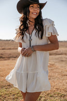 Woman in a white western boho dress, black cowboy hat, and bolo tie standing outdoors