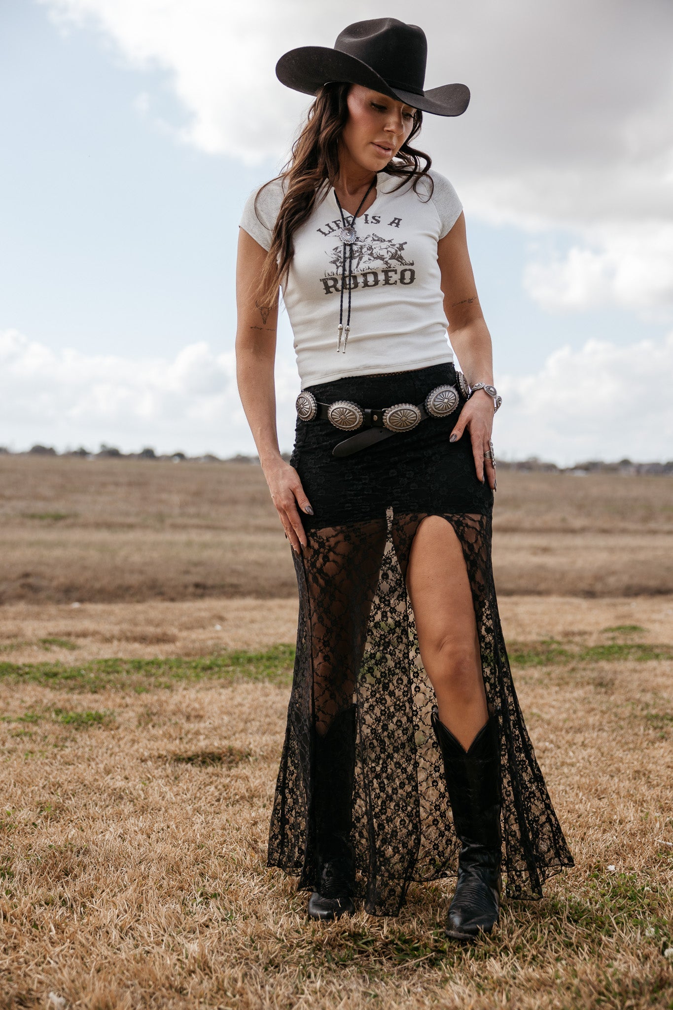 Woman in cowgirl hat, graphic tee, black lace skirt with slit, concho belt and western boots outdoors
