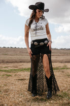 Woman in cowgirl hat, graphic tee, black lace skirt with slit, concho belt and western boots outdoors
