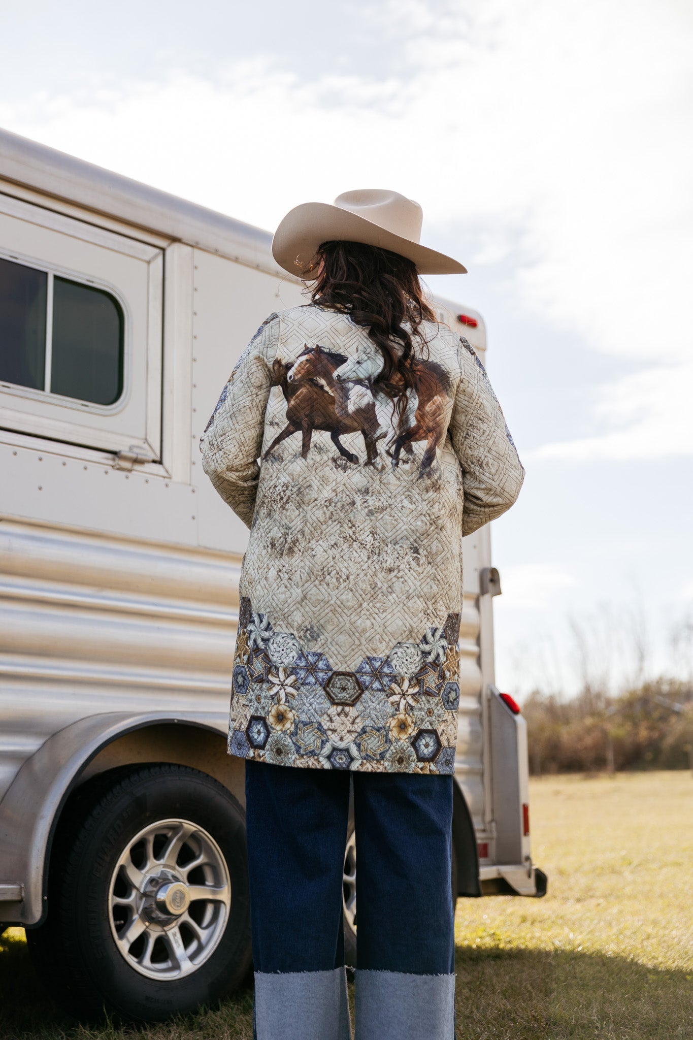 Woman in western boho coat with horse print, cowboy hat, standing by horse trailer outdoors