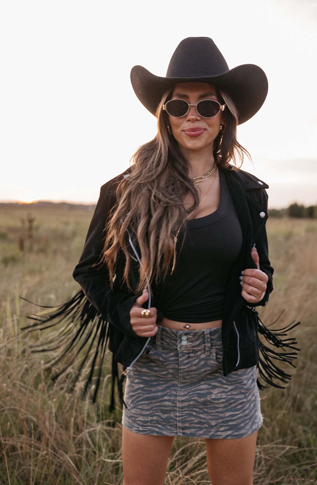 Woman wearing a black cowboy hat, fringe jacket, and sunglasses in a field.