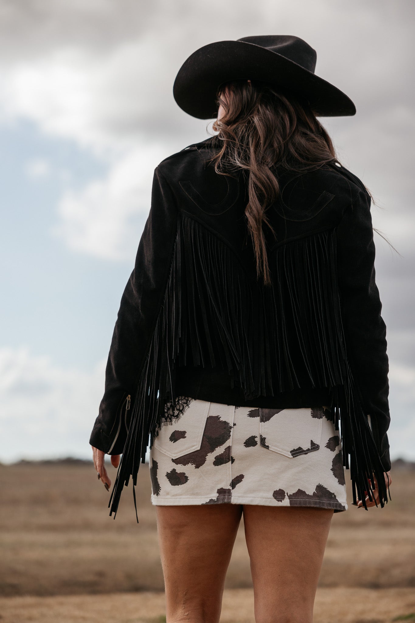 Woman in black fringe jacket and cow print skirt wearing cowboy hat in western landscape