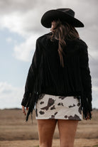Woman in black fringe jacket and cow print skirt wearing cowboy hat in western landscape