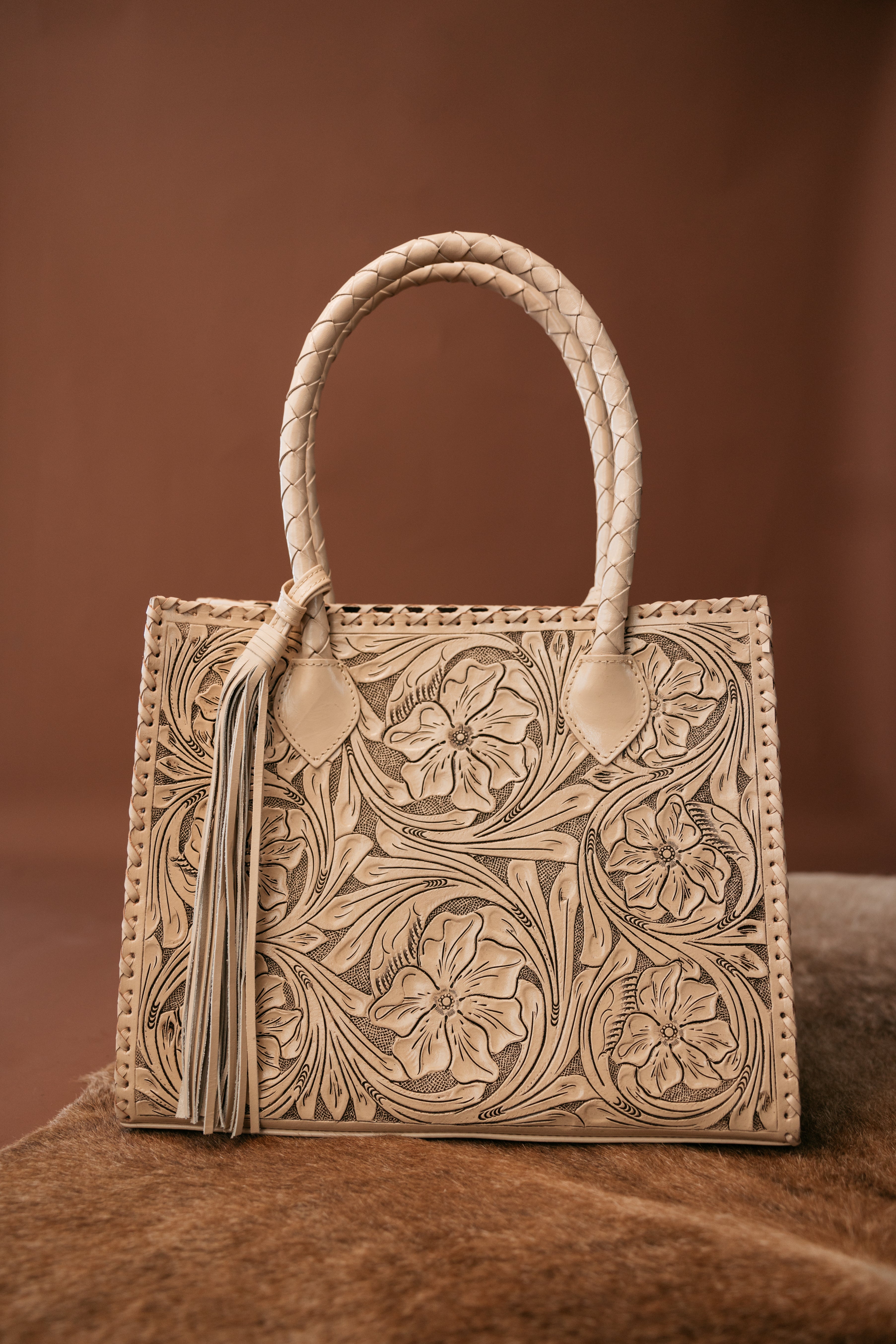 Western tooled leather handbag with floral design and braided handle on brown background