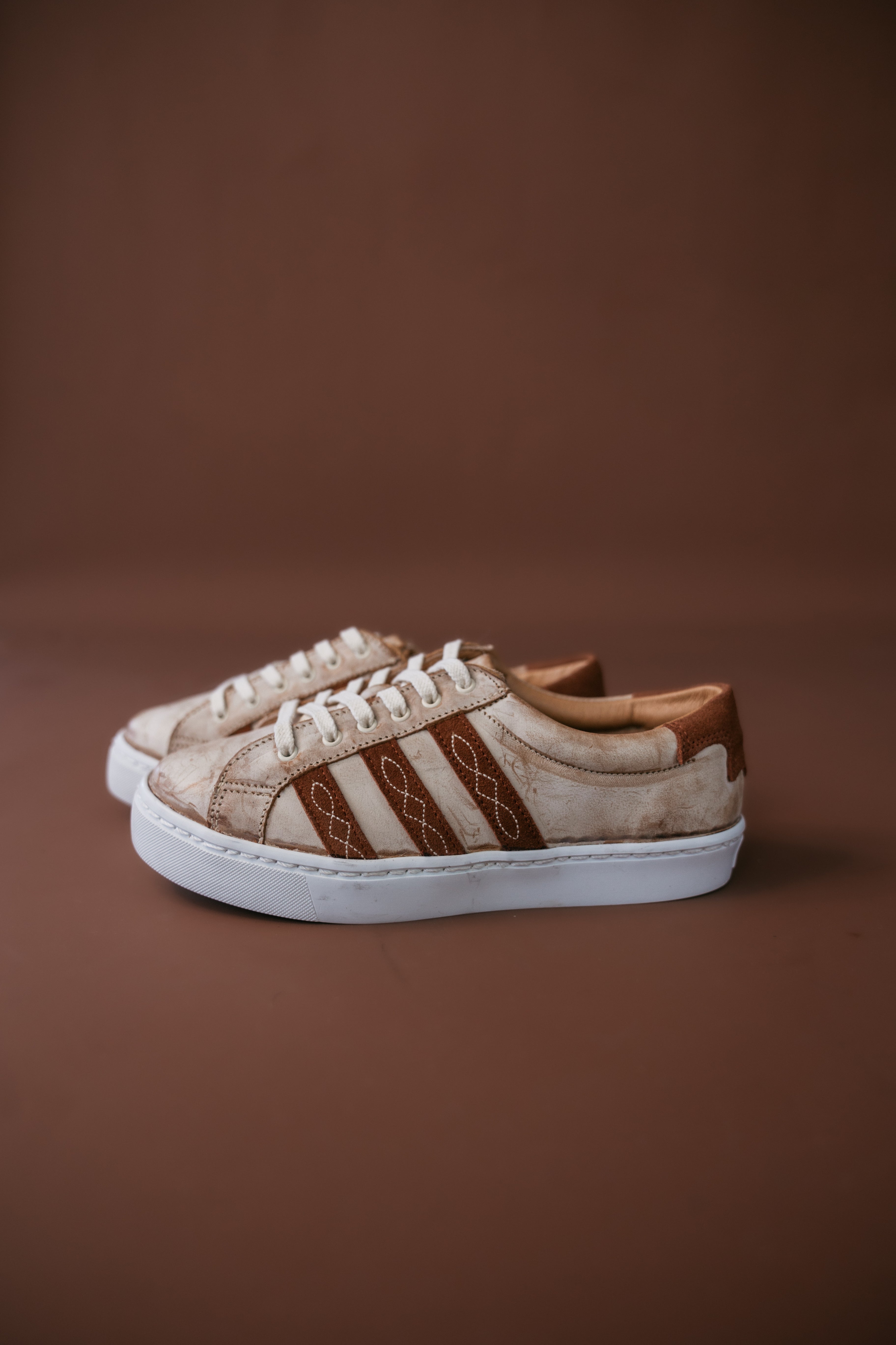 Beige and brown western sneakers with stitched detail, white sole, on brown background