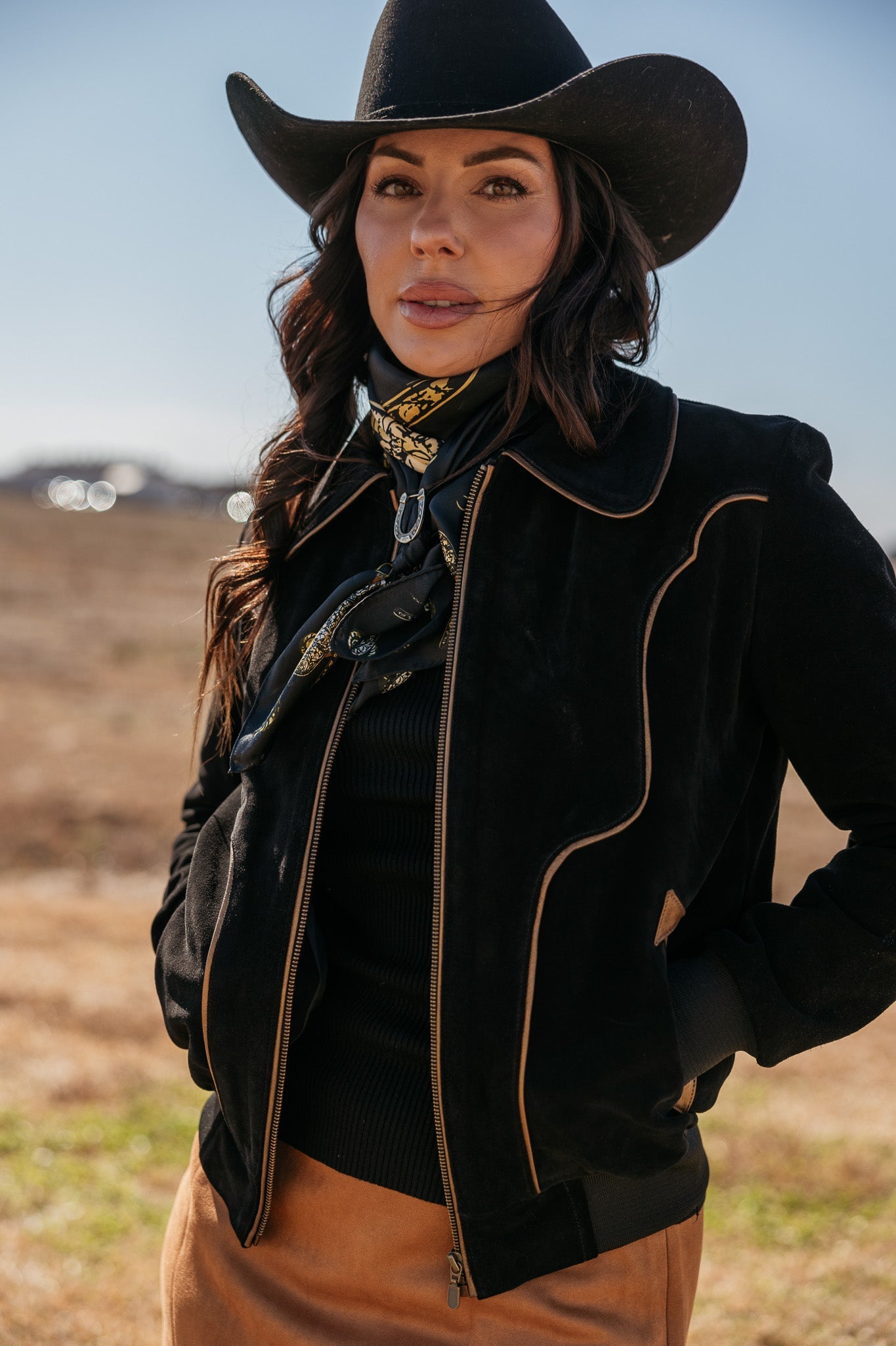 Woman in western boho outfit with black cowgirl hat, suede jacket, and bandana outdoors