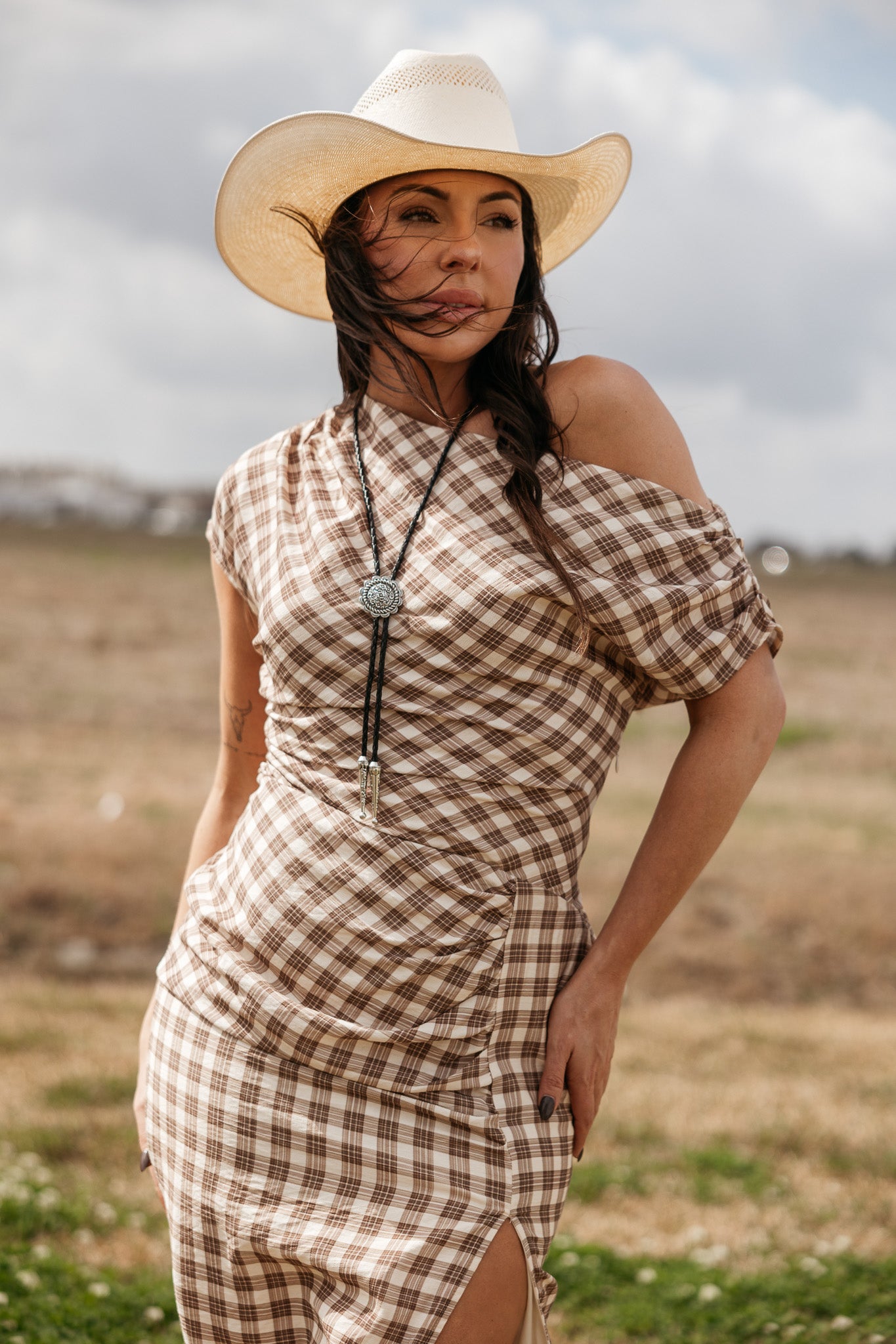 Woman in a brown plaid western dress, cowboy hat, and bolo tie standing outdoors