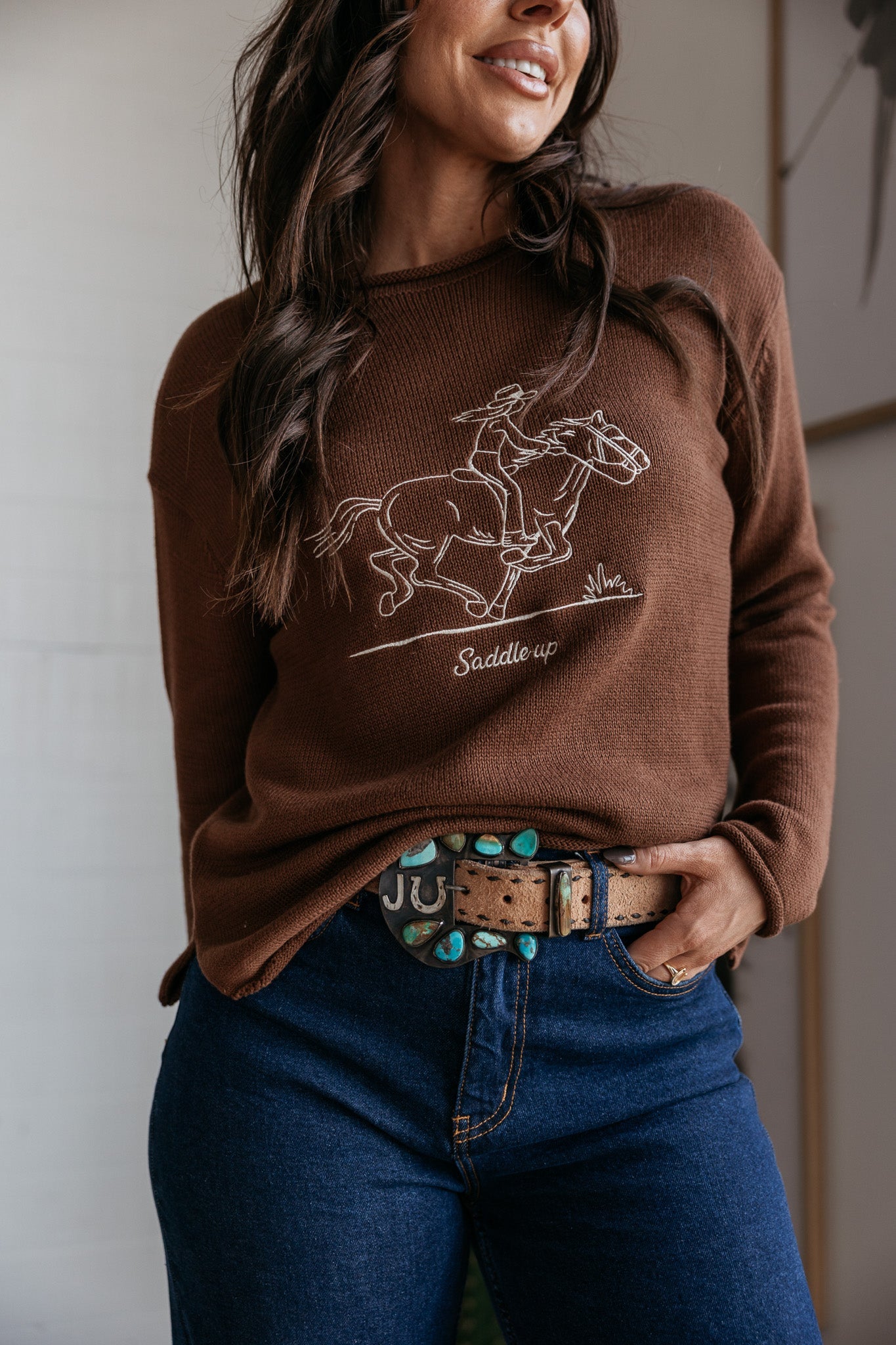Woman wearing brown 'saddle up' cowgirl graphic sweater, turquoise western belt, and blue jeans