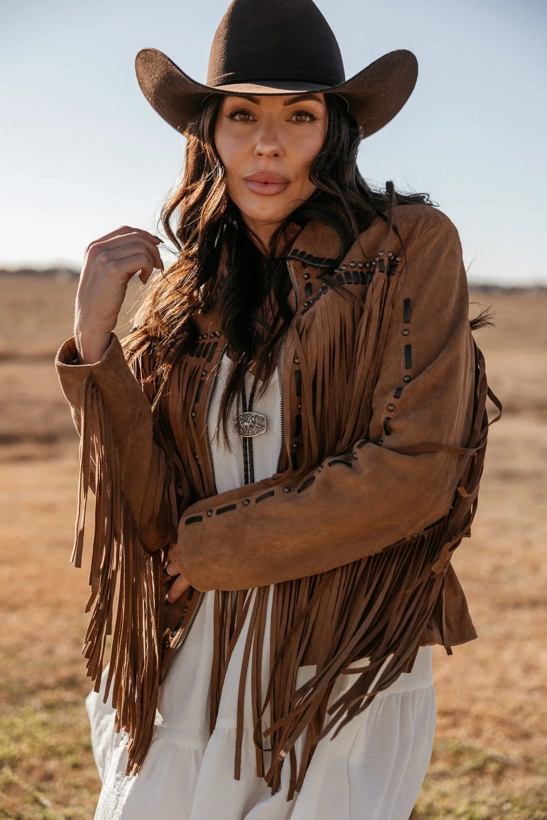 Woman in a brown fringe suede jacket and black cowboy hat in a western outdoor setting