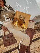 Gold western teardrop earrings on card, laid over cowgirl fashion magazine with boots and handbag.