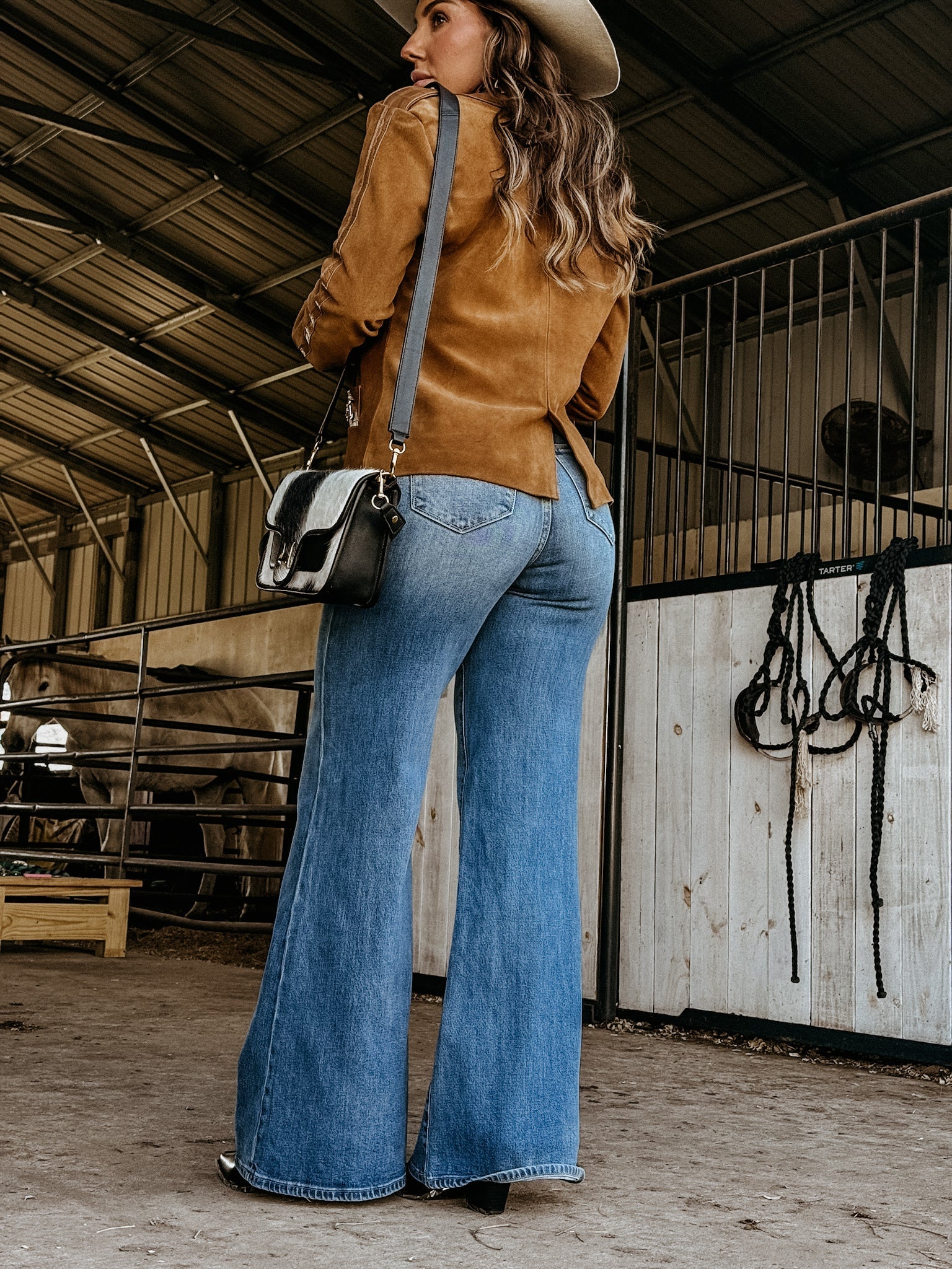 Woman in a suede jacket, flared jeans, and cowboy hat in a western barn setting