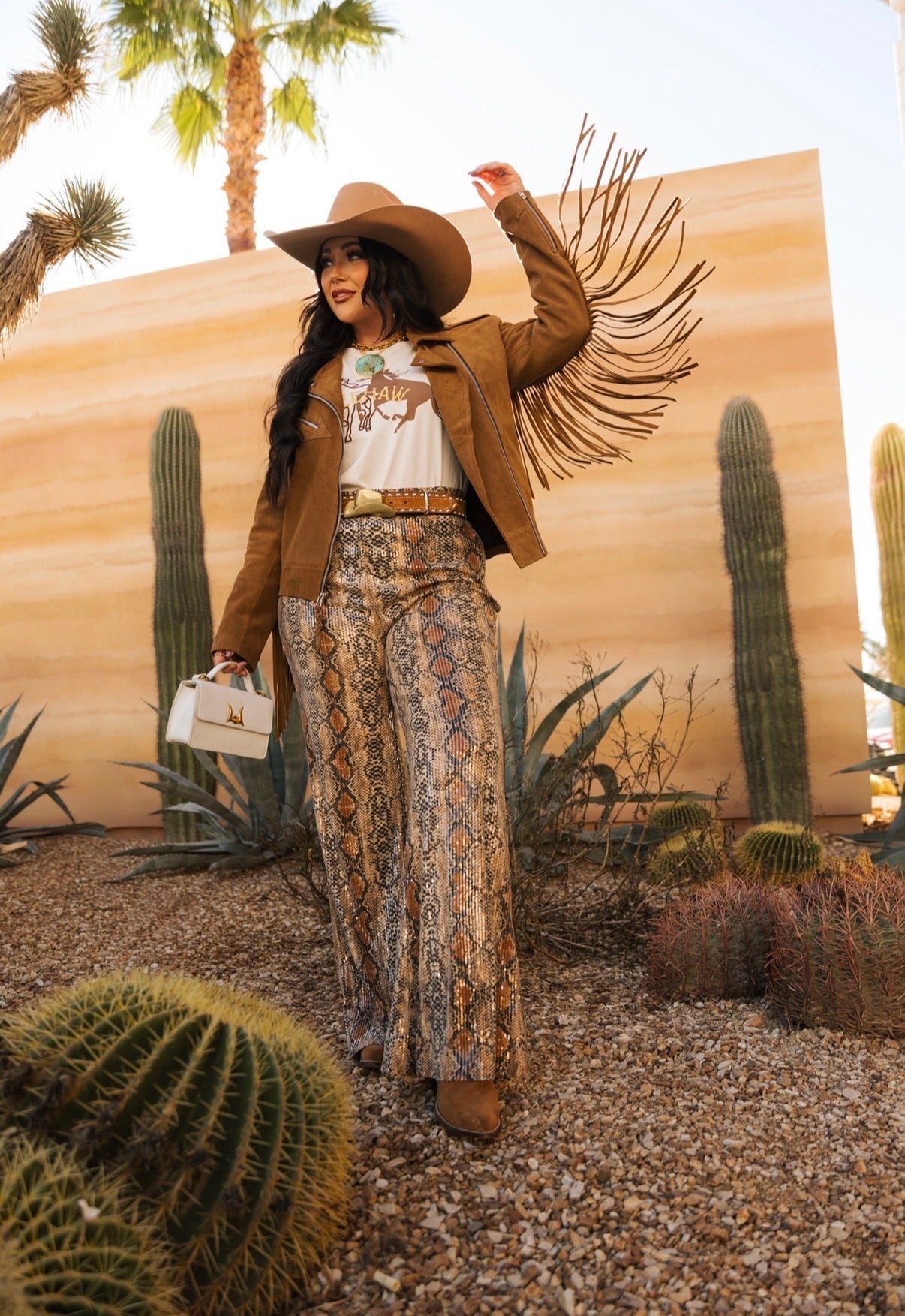Woman in western boho outfit with fringe jacket, cowboy hat, snake print pants, and boots outdoors