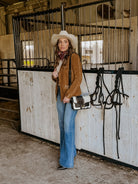 Woman in western boho outfit with suede jacket, cowboy hat, flared jeans in a horse stable