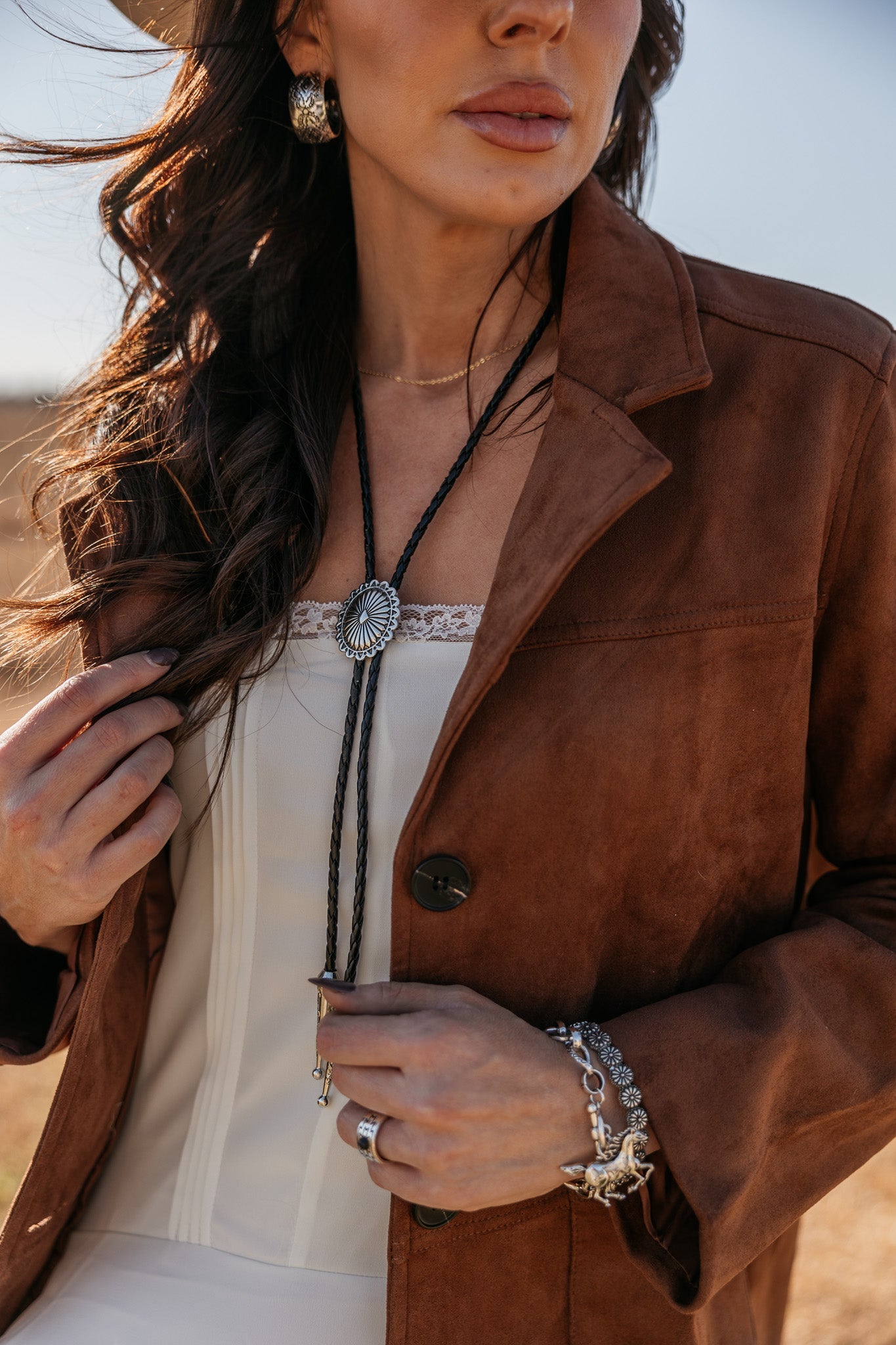 Woman in brown suede jacket, western bolo tie, white lace top, silver jewelry outdoors