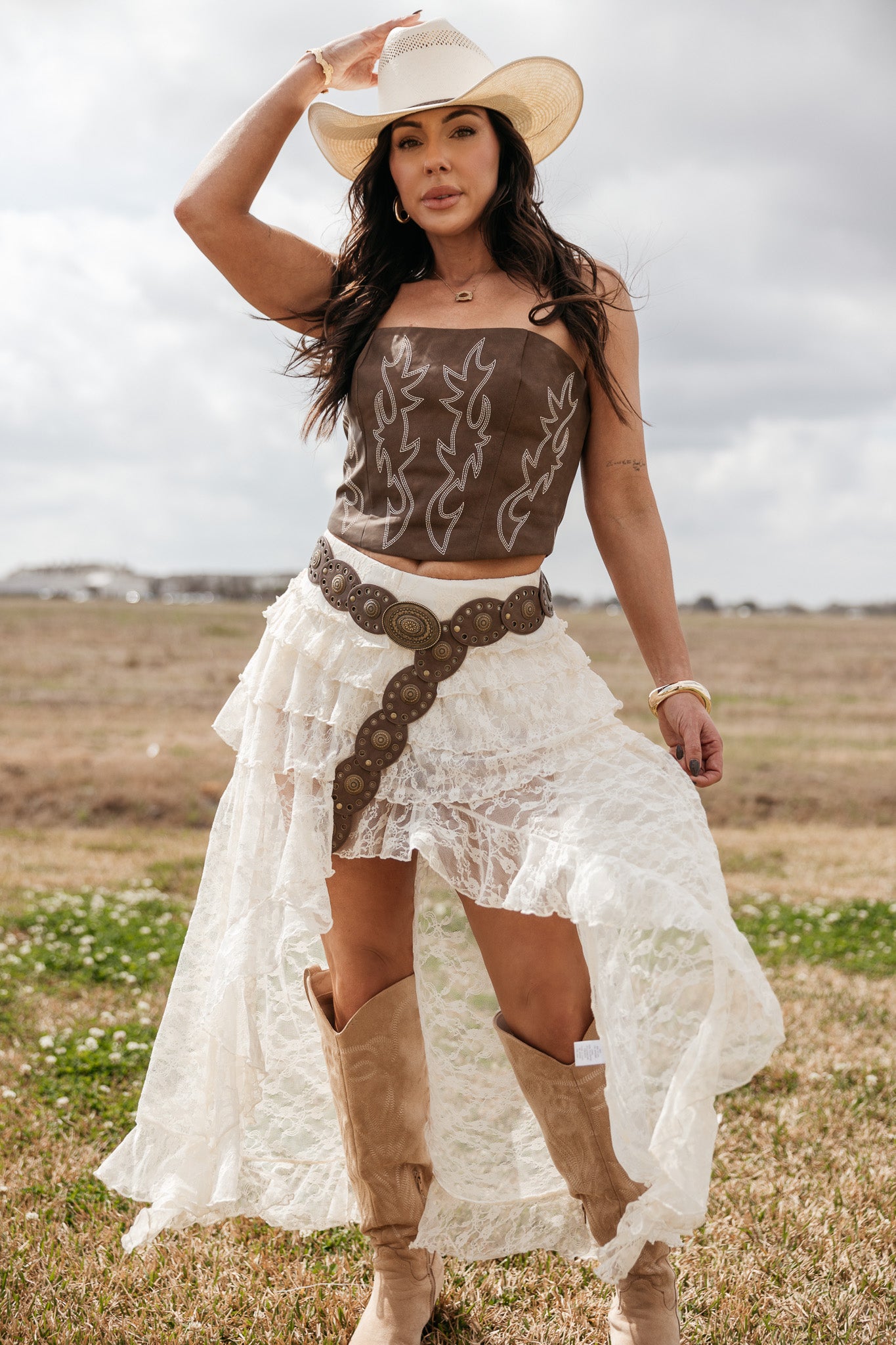 Woman in western boho outfit with lace skirt, brown corset top, cowboy boots, and hat outdoors