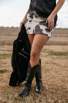 Woman in a cow print mini skirt, black lace top, black western boots, holding suede jacket outdoors