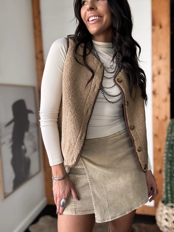 Woman wearing a beige vest over a white long-sleeve shirt and tan skirt indoors.