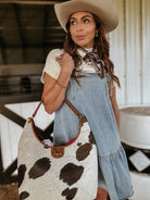 Woman in western boho outfit with cowboy hat, denim dress, and cow print bag