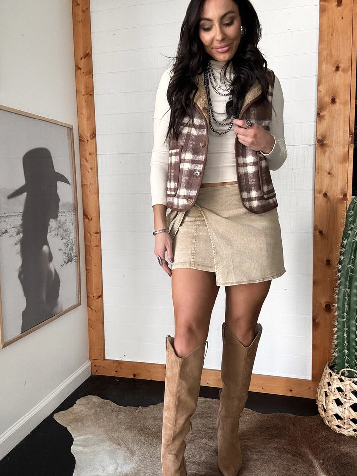 Woman wearing a plaid vest, beige skirt, and brown knee-high boots indoors.