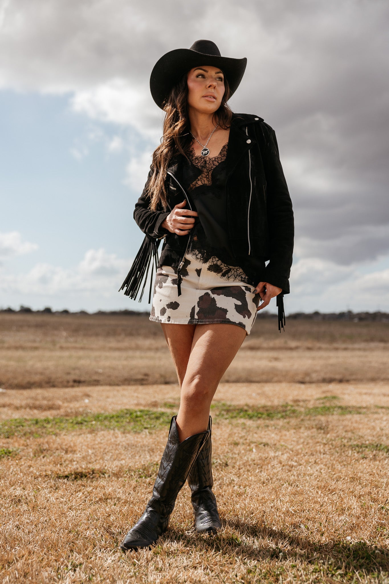 Woman in black cowboy hat, lace top, fringe jacket, cow print skirt, and western boots outdoors