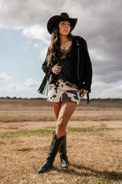 Woman in black cowboy hat, lace top, fringe jacket, cow print skirt, and western boots outdoors