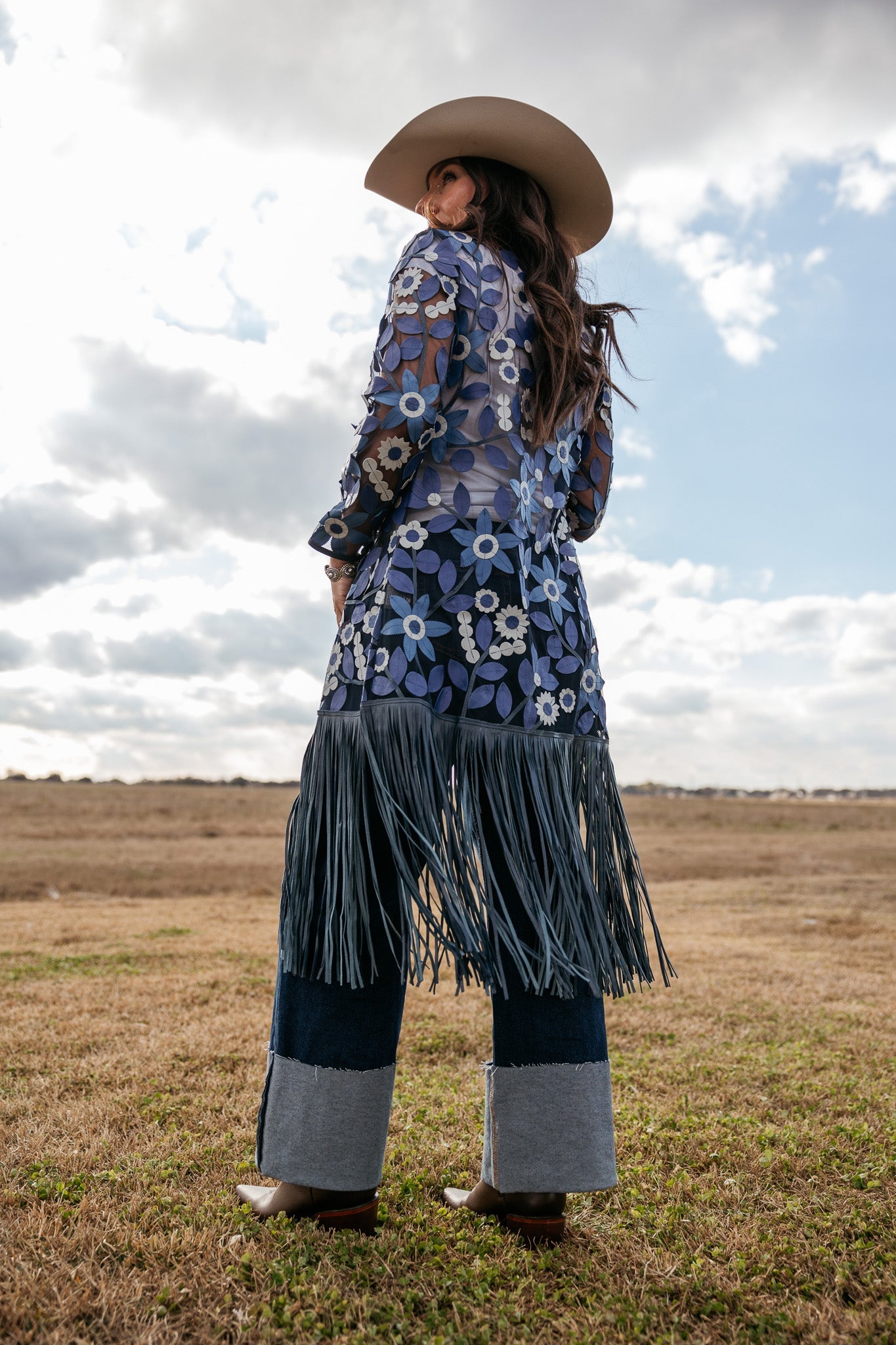 Woman in western boho outfit with floral sheer top, fringe skirt, wide cuff jeans, and cowboy hat standing outdoors