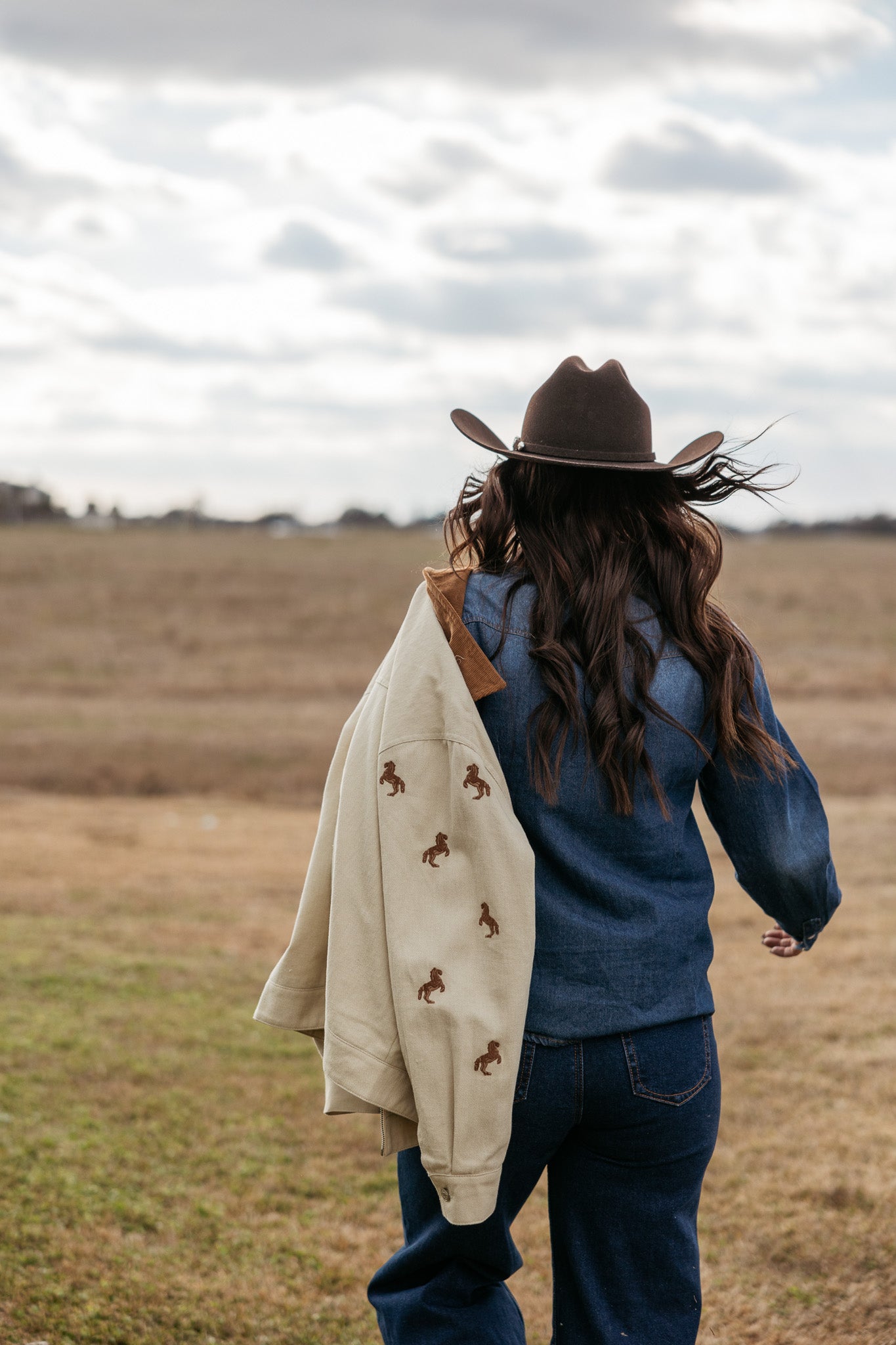 Woman in cowboy hat and denim outfit outdoors, holding tan jacket with horse embroidery, western style