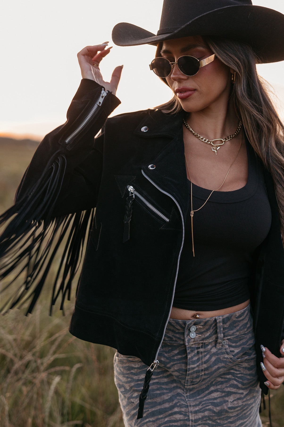 Woman in a black fringe jacket, black top, and denim skirt with a cowboy hat in a field.