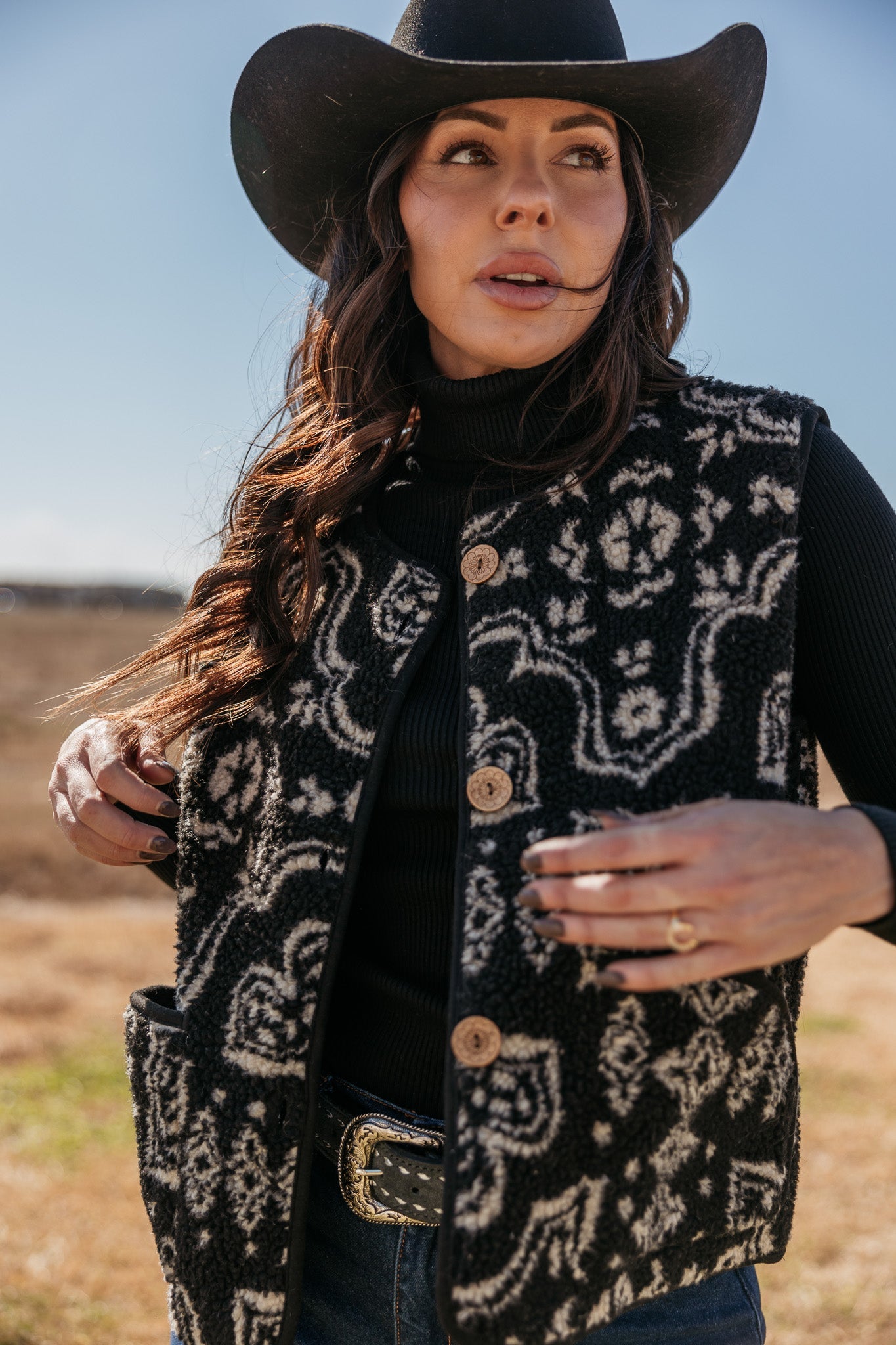 Woman in western black felt hat, patterned sherpa vest, and belt, outdoors in sunlight