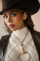 Woman in cowgirl hat, cream sweater, leather vest, with Western jewelry and horse earrings