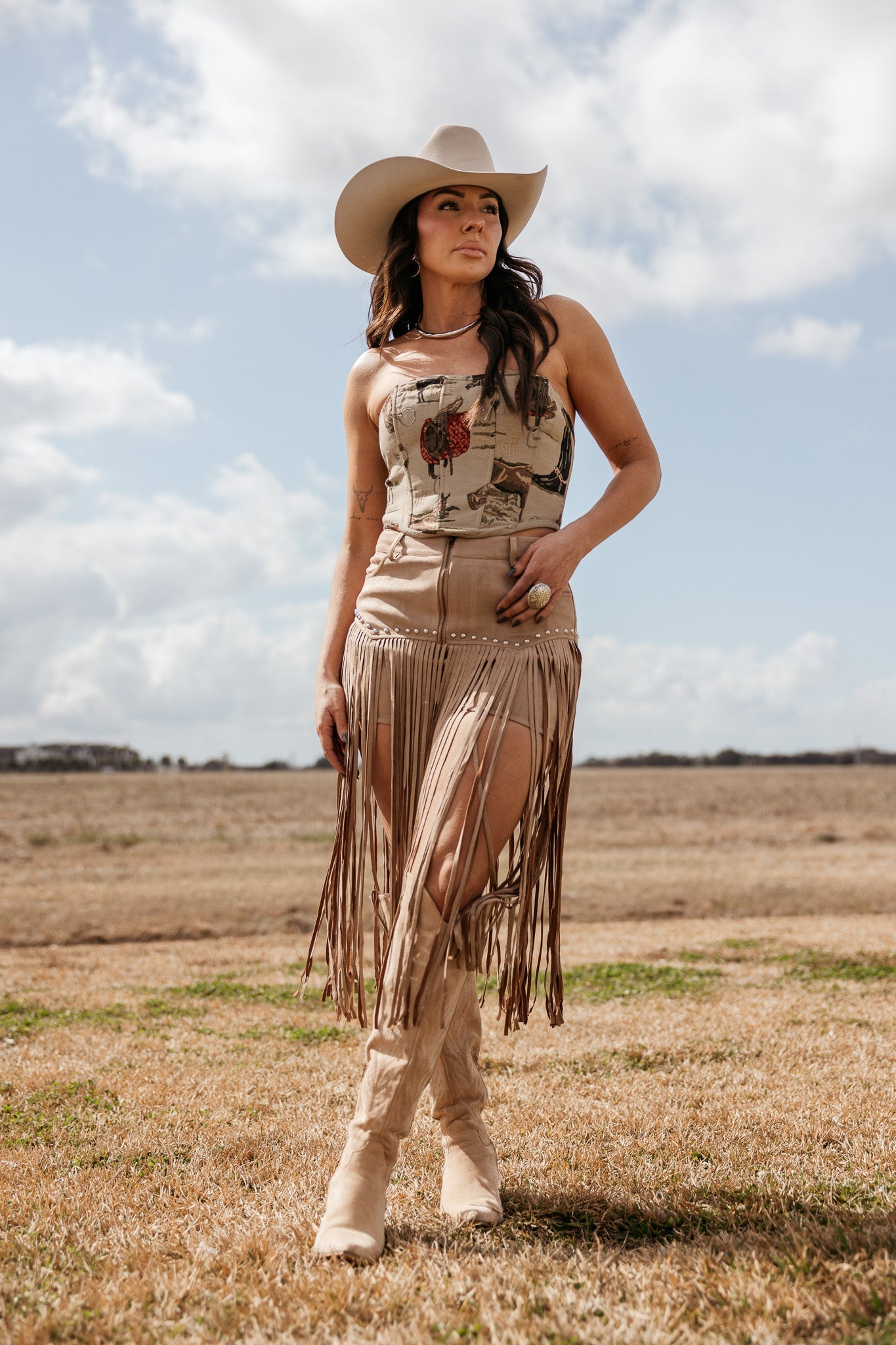 Woman in western boho outfit with beige cowboy hat, fringe skirt, and boots standing outdoors