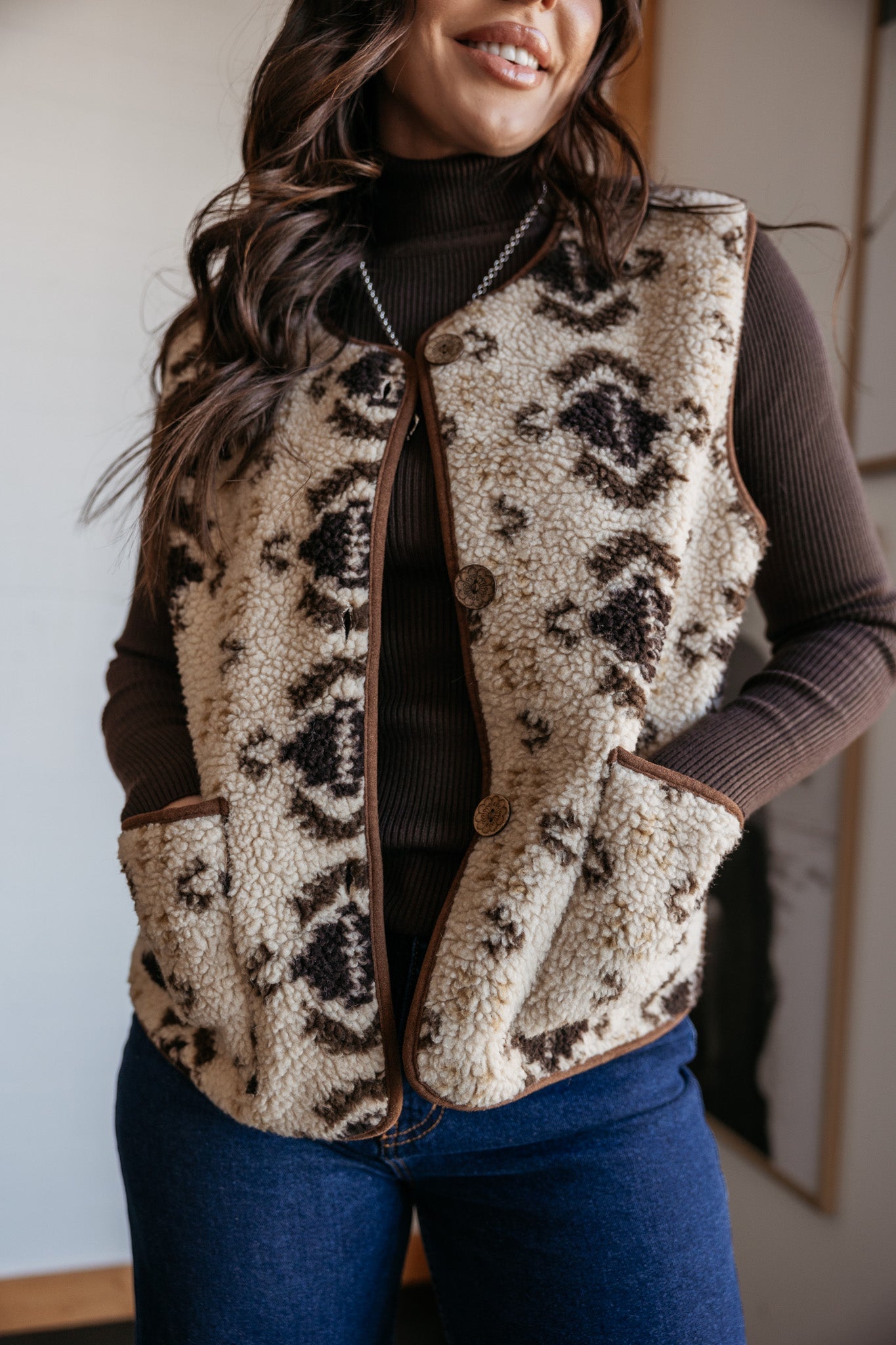 Woman wearing a western sherpa vest with Aztec pattern, brown turtleneck, and blue jeans.