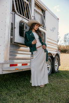 Woman in cowboy hat, green suede jacket, and cream maxi skirt leaning on horse trailer, western outfit