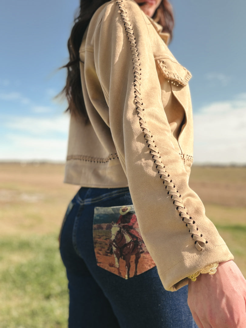 Person wearing a beige jacket with lace details and blue jeans with a horse print pocket in an outdoor setting.