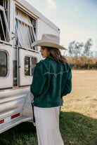 Woman in a wide-brim hat and emerald western jacket by a horse trailer, outdoor boho style