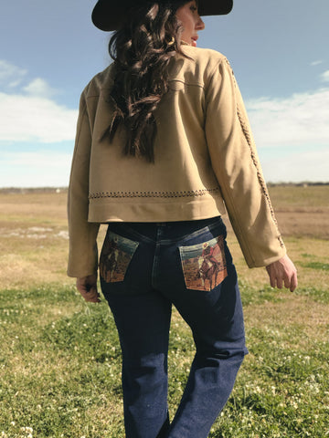 Person wearing a beige jacket and blue jeans with patterned pockets in an open field.