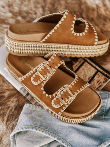 Camel platform sandals featuring two stitched buckle straps with gold adjustable hardware, a braided raffia-style platform, textured sole for added grip, and a soft cushioned insole for comfort.