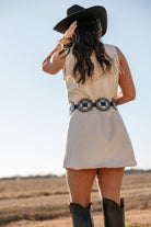 Woman in cream fringe dress, concho belt, black cowgirl hat and boots in a western field