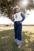 Woman in a cowboy hat, wide-leg jeans, white knit vest and boots, posing outdoors in a western ranch setting