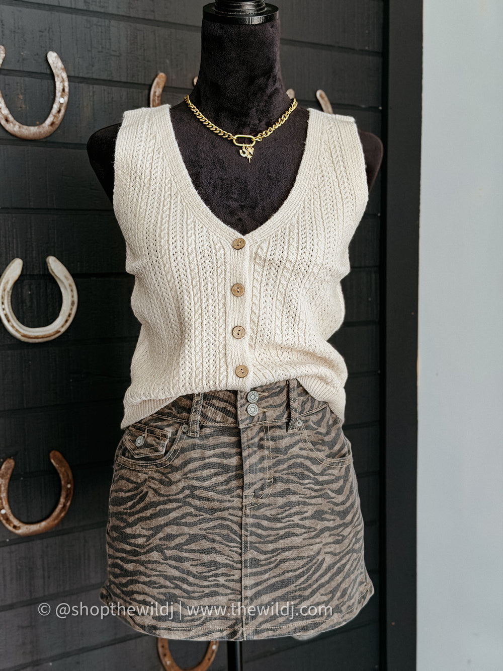 Mannequin wearing a cream knitted vest and zebra print skirt against a dark wooden wall with horseshoes.