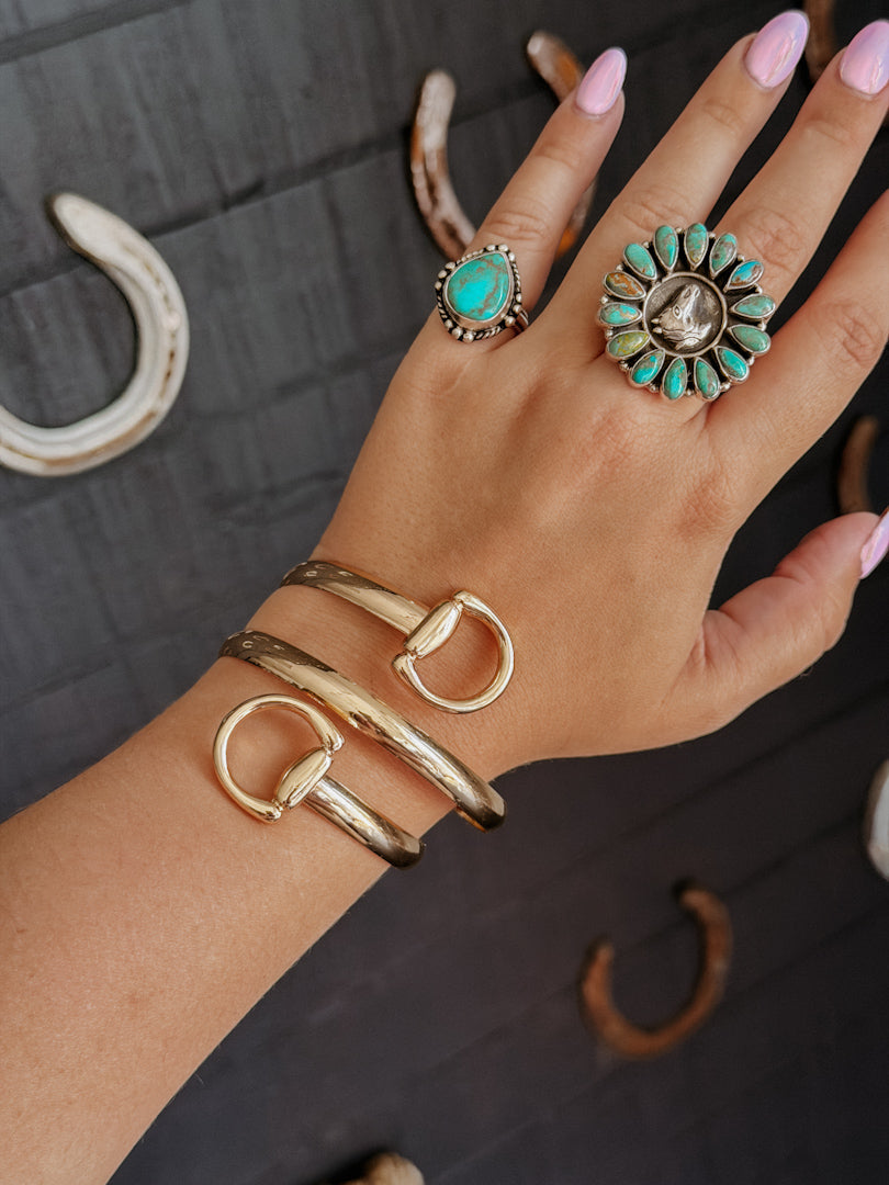 Hand wearing gold bracelet and turquoise rings on a dark wooden surface