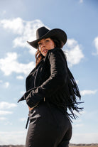 Woman in black fringe jacket and cowboy hat posing outdoors under blue sky, western fashion