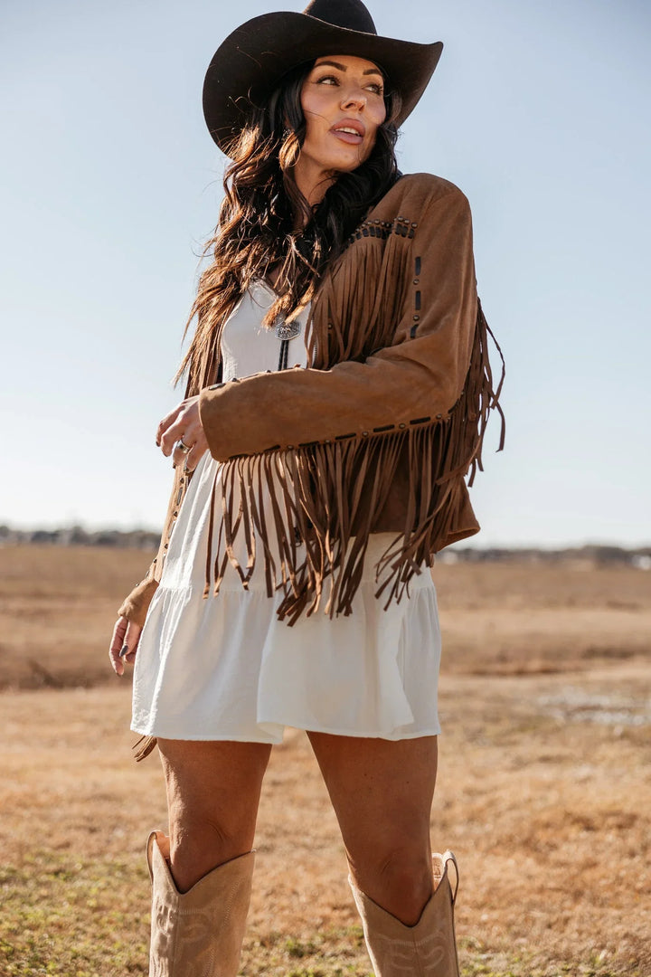 Woman in western cowgirl outfit with suede fringe jacket, white dress, cowboy hat and boots