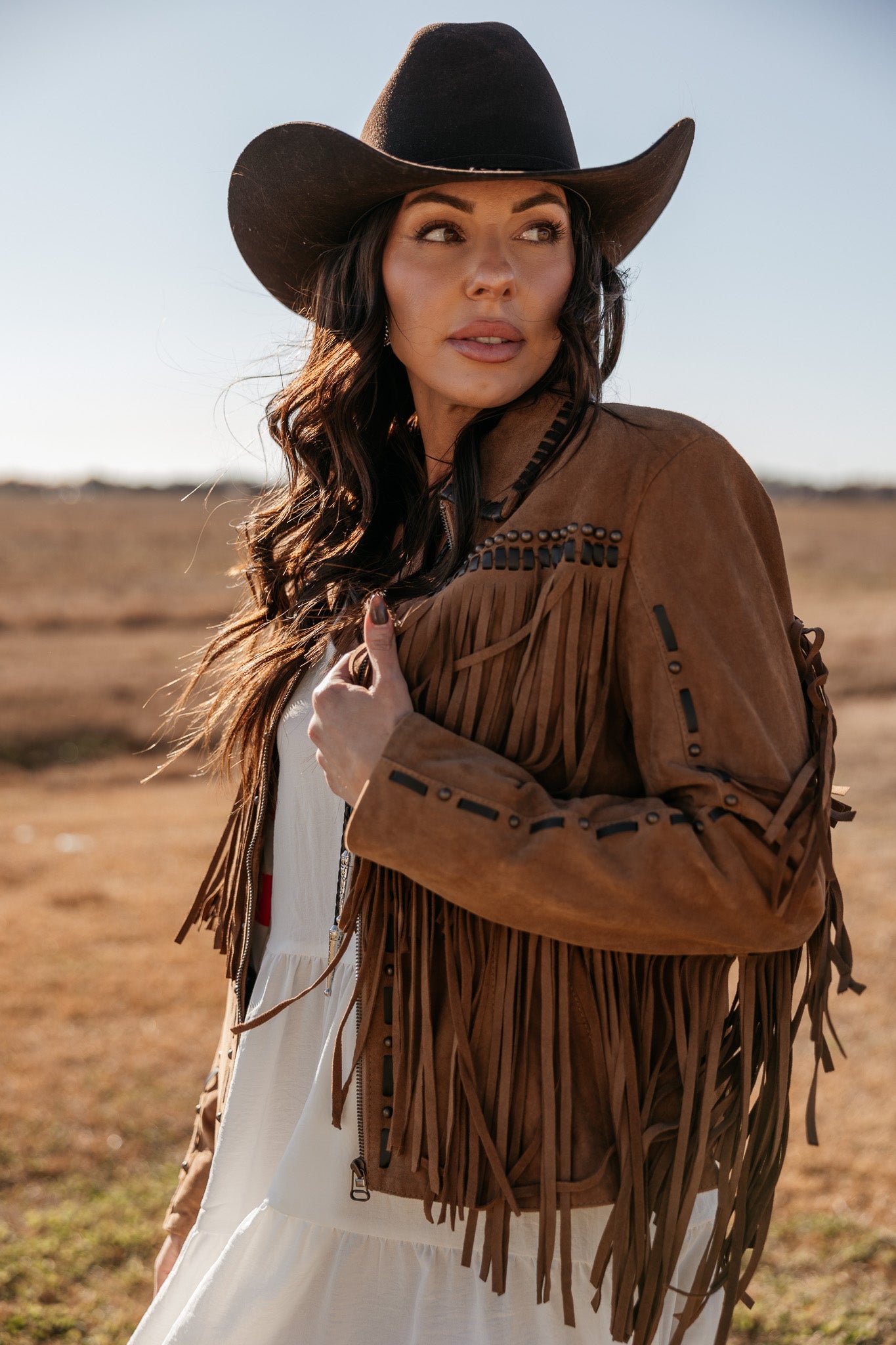 Woman in a brown fringe suede jacket and black cowboy hat in a western outdoor setting