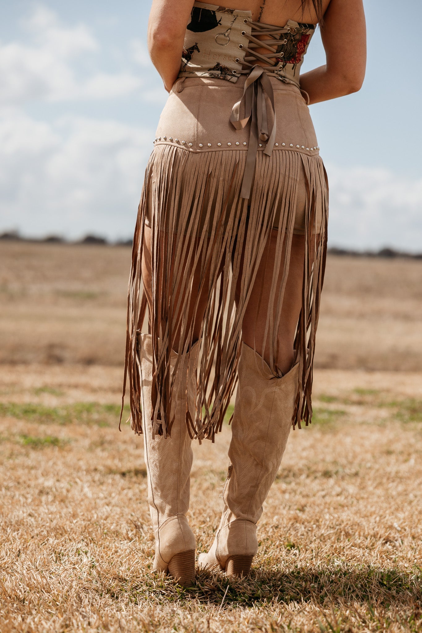 Woman wearing tan fringe western skirt, corset top, and cowboy boots outdoors