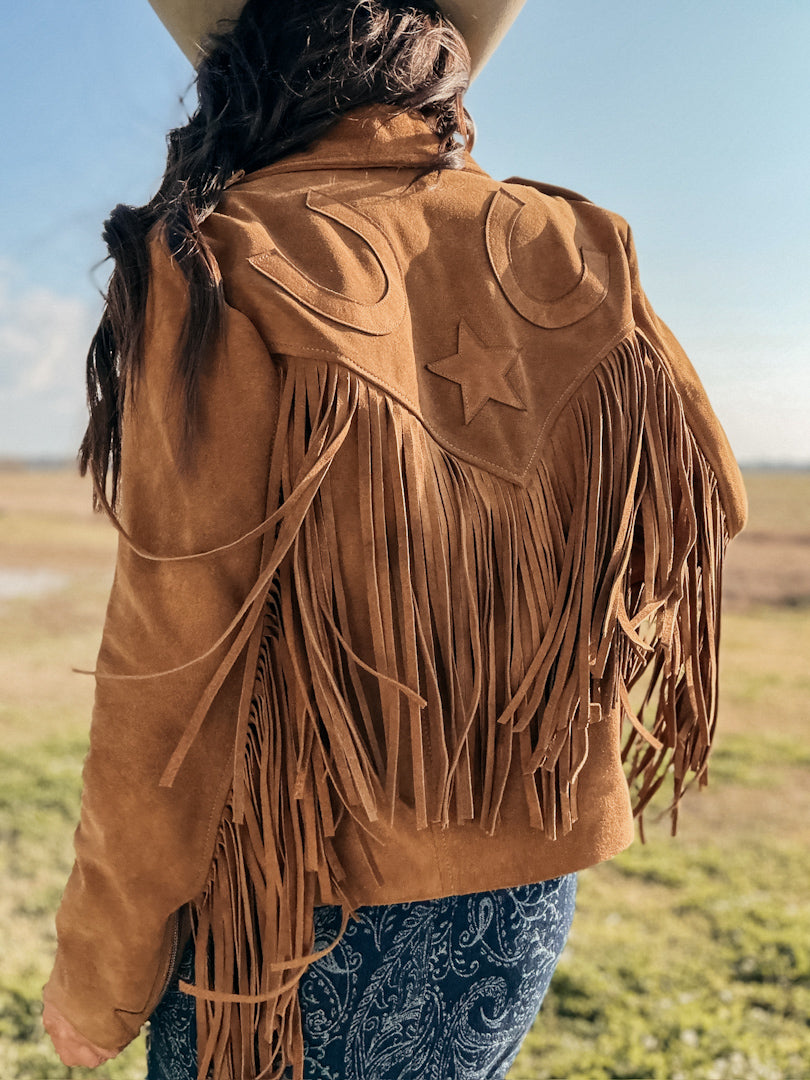 Woman in suede fringe western jacket with star and horseshoe details, cowboy hat, and paisley jeans outdoors.