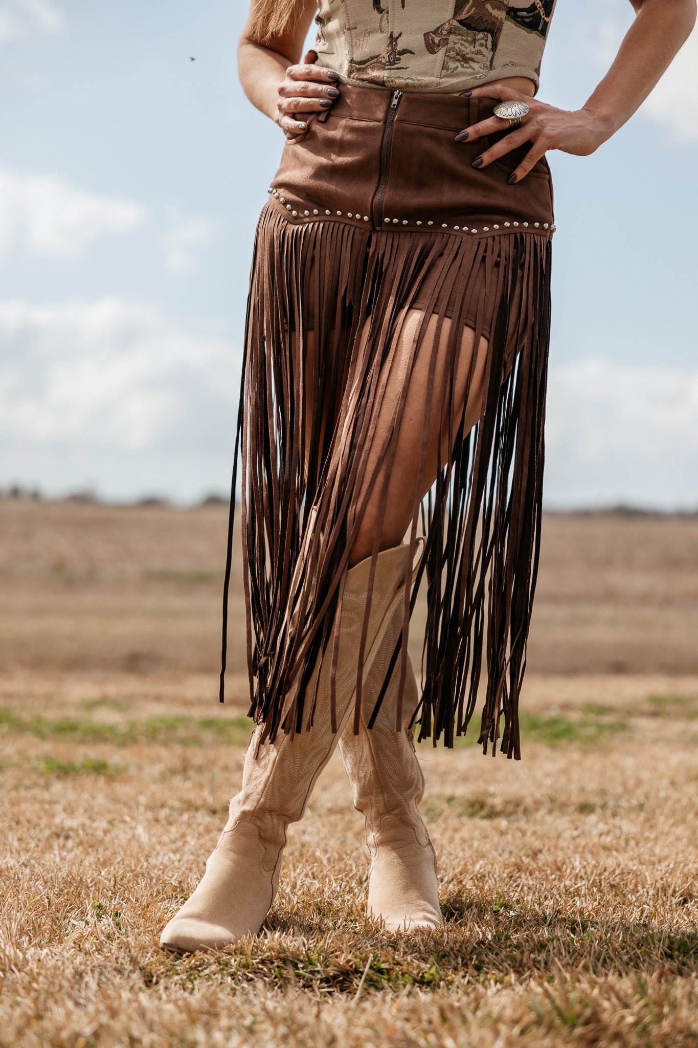 Woman wearing brown fringe suede skirt, western graphic top, and tan cowgirl boots outdoors